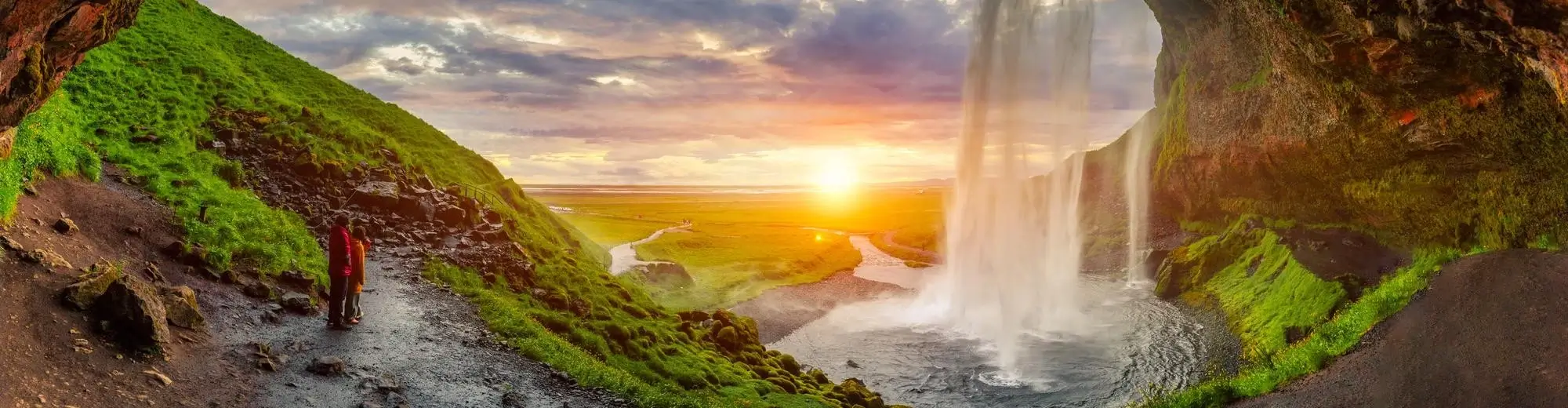 person on a scenic path behind a large waterfall at sunset.