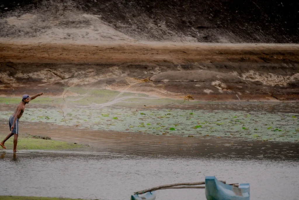 a fisherman casting a net in a river with a small boat.